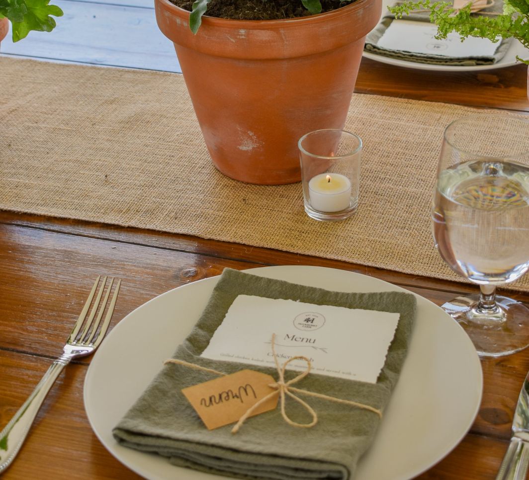 A place setting with a sage napkin, handwritten name tag and menu card beneath a potted fern