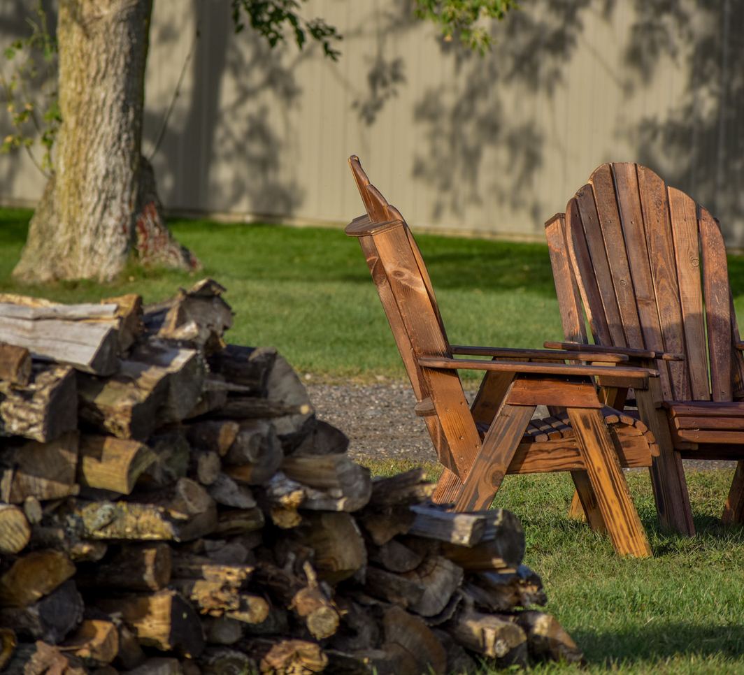 Wooden Adirondack chairs beside a stacked woodpile under a shade tree